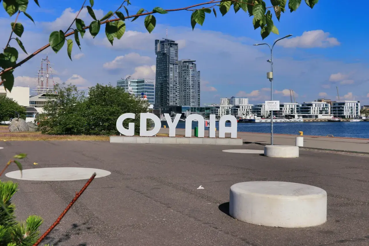 Napis GDYNIA na tle nowoczesnej architektury i żaglowca. Widok z Gdynia Rynek Główny.
