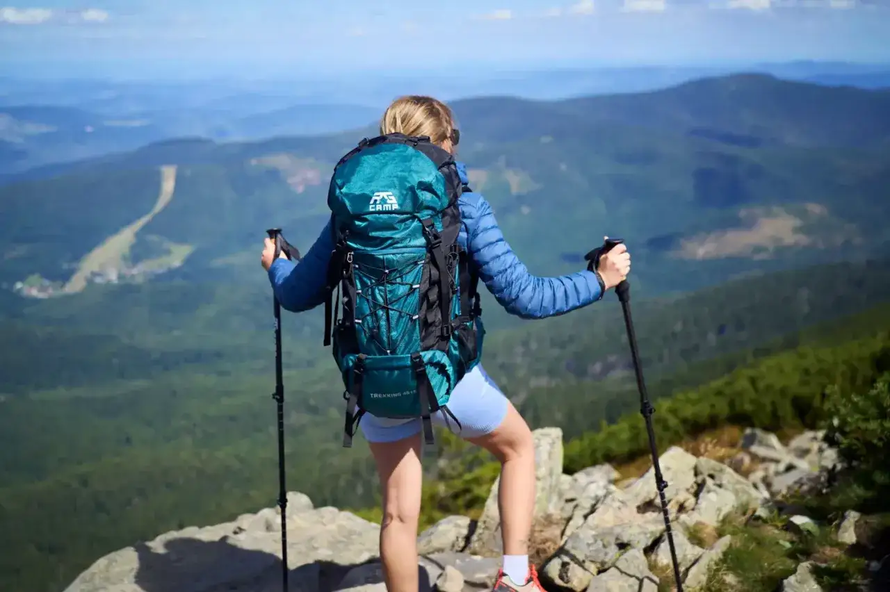 Kobieta z turkusowym plecakiem na jednodniową wycieczkę w góry, z kijkami trekkingowymi, podziwia panoramę.