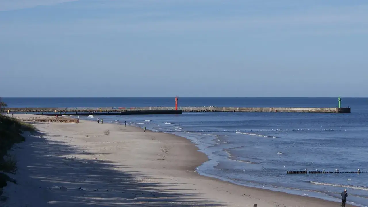 Plaża w Mrzeżynie z falochronem i latarniami morskimi. Spacerowicze cieszą się spokojem, a fale delikatnie rozbijają się o brzeg. To idealne miejsce na relaks i odkrywanie mrzeżyno atrakcje.