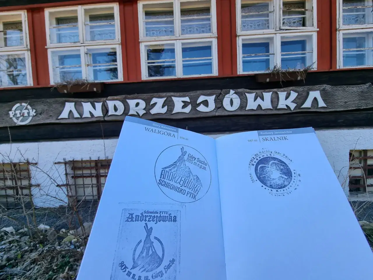 Przed schroniskiem Andrzejówka, z widokiem na budynek i otwartą książkę z pieczątkami z Waligóry i Skalnika.