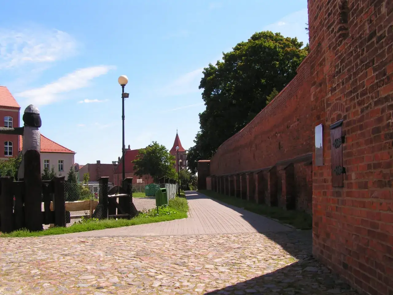 Spacer po Chełmnie: brukowana ścieżka wzdłuż ceglanych murów obronnych, z widokiem na zabytkowe budynki i wieżę kościelną. Atrakcje miasta czekają!