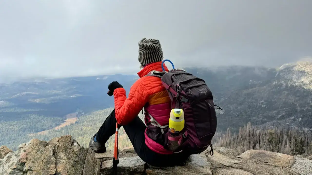 Osoba z plecakiem i kijami siedzi na skale, podziwiając widok. To idealne miejsce na trekking vs hiking, gdzie można docenić piękno natury.