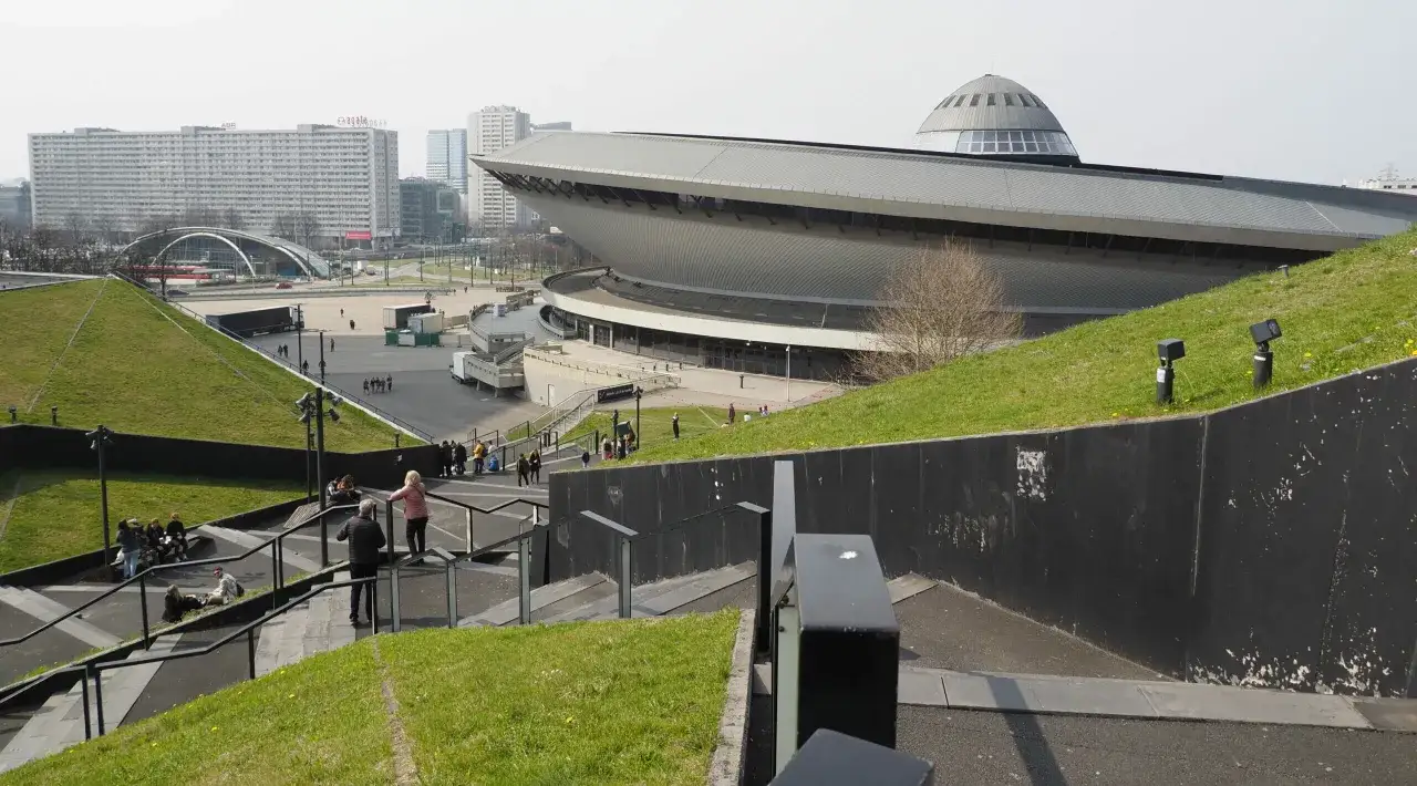 Katowice atrakcje: Spodek i Międzynarodowe Centrum Kongresowe na tle bloków mieszkalnych. Ludzie spacerują po zielonych terenach i schodach.