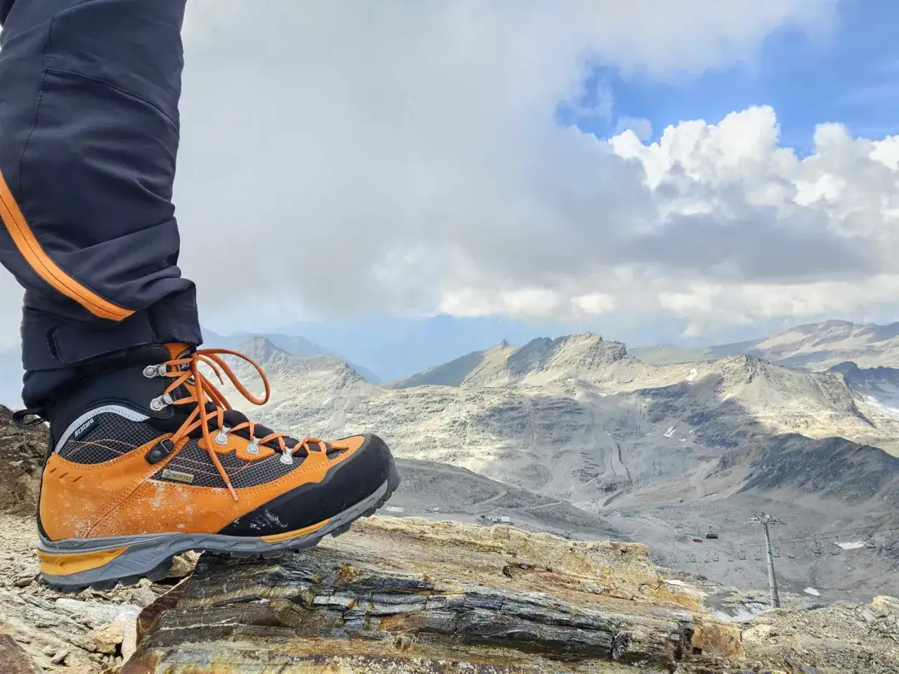 Pomarańczowy but trekkingowy na skale, w tle góry i kolejka linowa. Czy to buty firmy Rab?