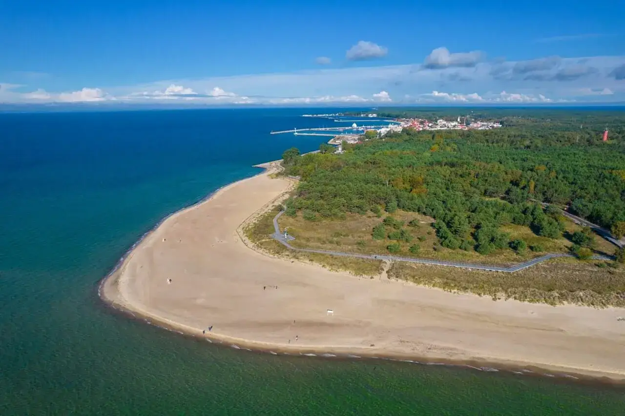 Plaża i las na Helu. Widok z lotu ptaka na turkusowe morze, piaszczystą plażę i zielony las. W oddali widać zabudowania i port. Hel atrakcje czekają!