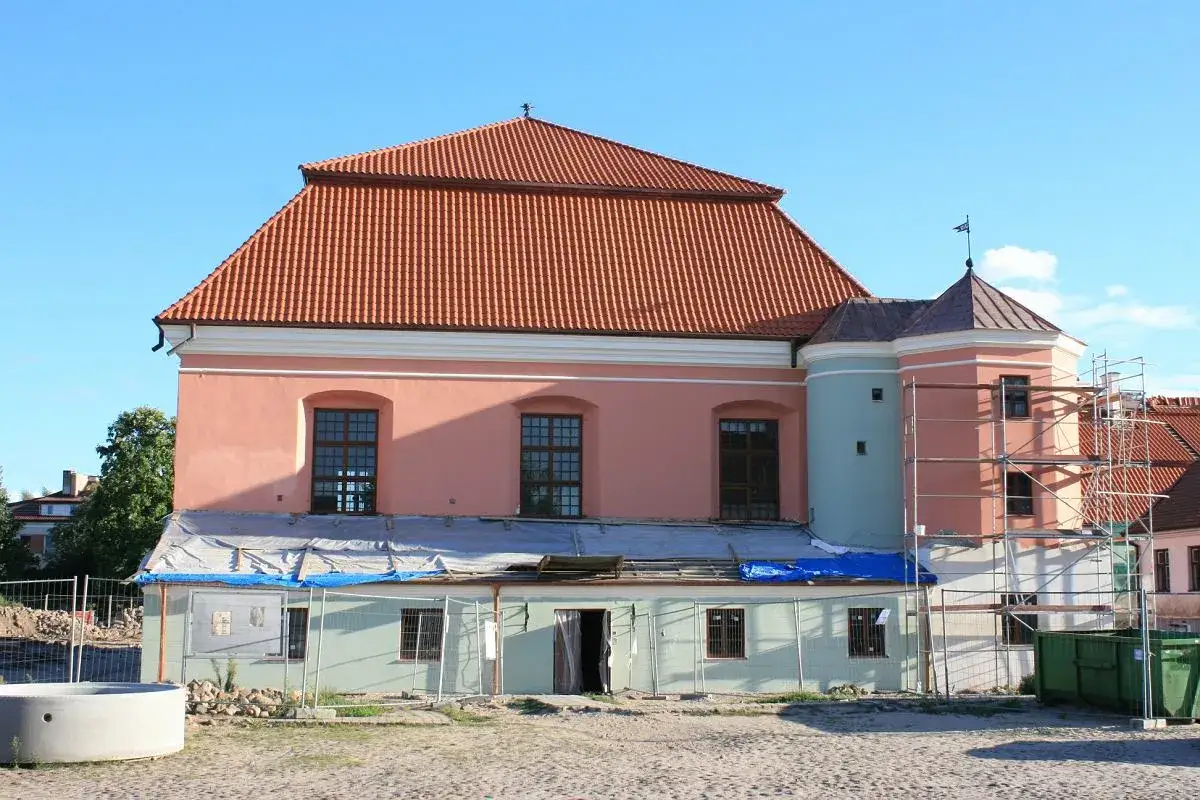 Różowy budynek z niebieskim fragmentem i rusztowaniami, w tle drzewa i niebo. To jedna z tykocin atrakcji w trakcie renowacji.