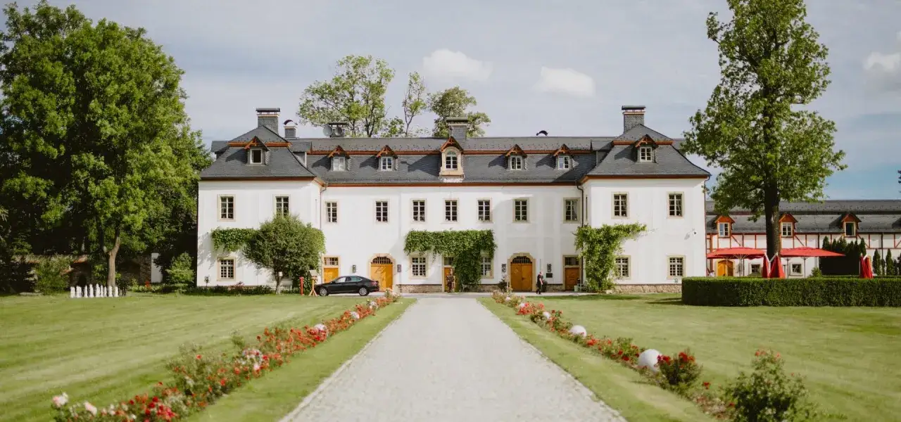 Malowniczy dwór z białej fasady, otoczony zielonym ogrodem i kwitnącymi rabatami. To idealne miejsce na spacer po dolinie pałaców i ogrodów.
