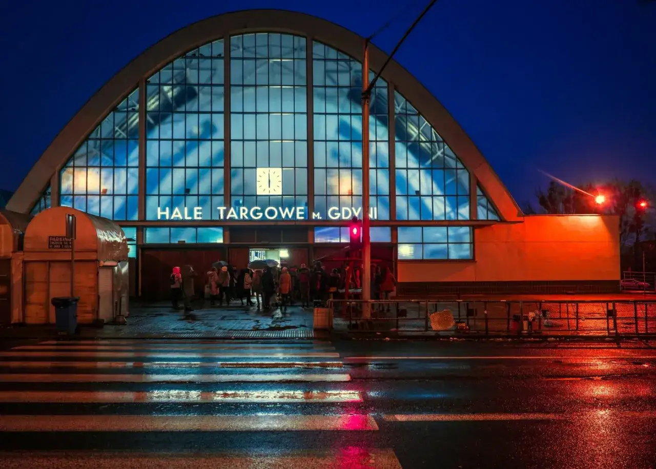 Wieczór na gdynia rynek główny. Ludzie z parasolami czekają przed Halą Targową, mokry asfalt odbija światła.