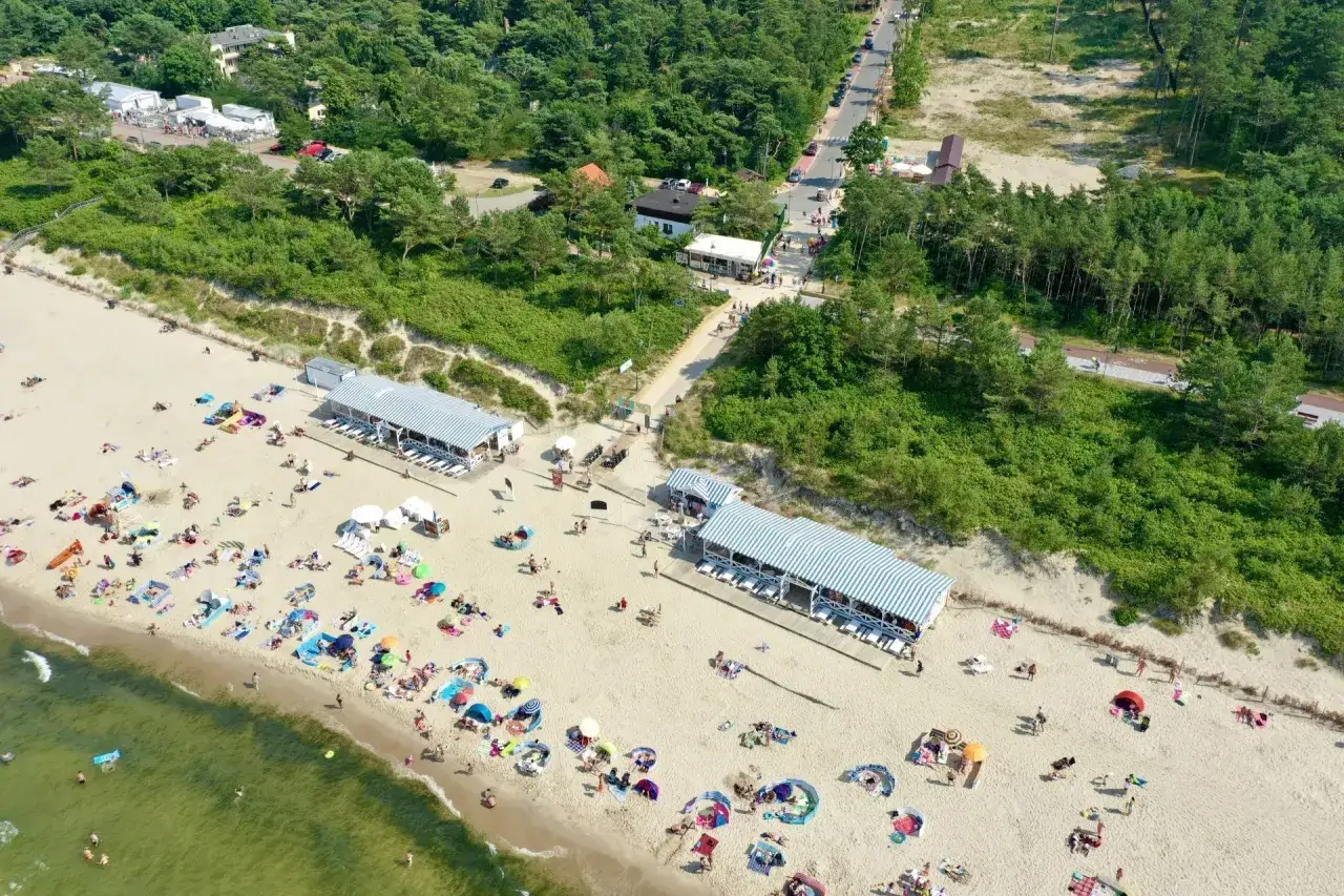 Plaża w Krynicy Morskiej pełna plażowiczów, parasoli i budynków z pasiatymi dachami. W tle las i droga z samochodami. Krynica Morska atrakcje dla każdego.