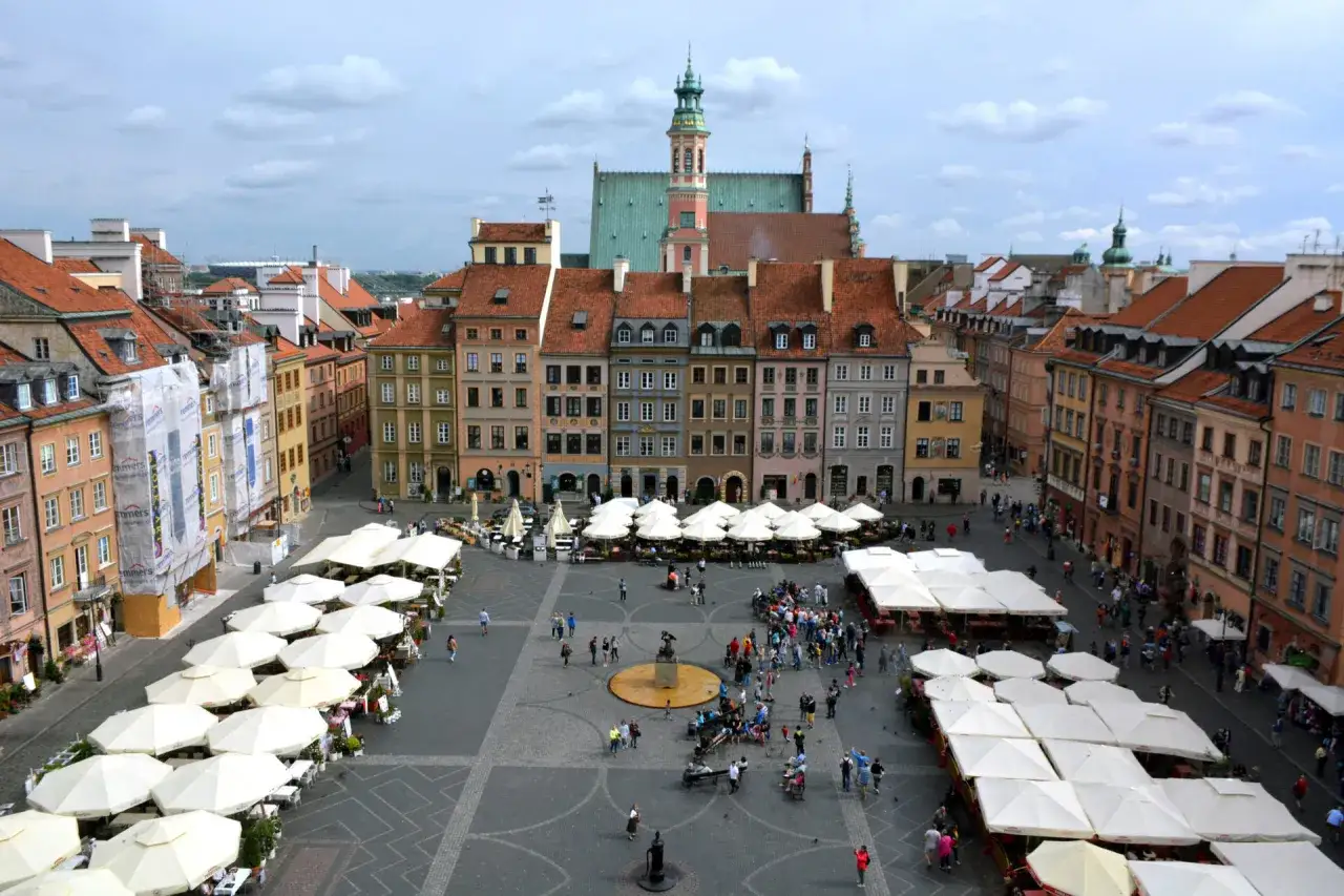 Rynek Starego Miasta w Warszawie, polskie Carcassonne, tętni życiem. Ludzie spacerują, siedzą w ogródkach kawiarnianych pod białymi parasolami.