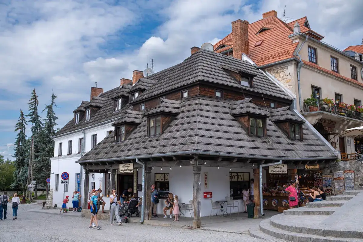Urokliwy rynek w Kazimierzu Dolnym z kamieniczkami i drewnianymi dachami. Ludzie spacerują, kupują lody i pamiątki.
